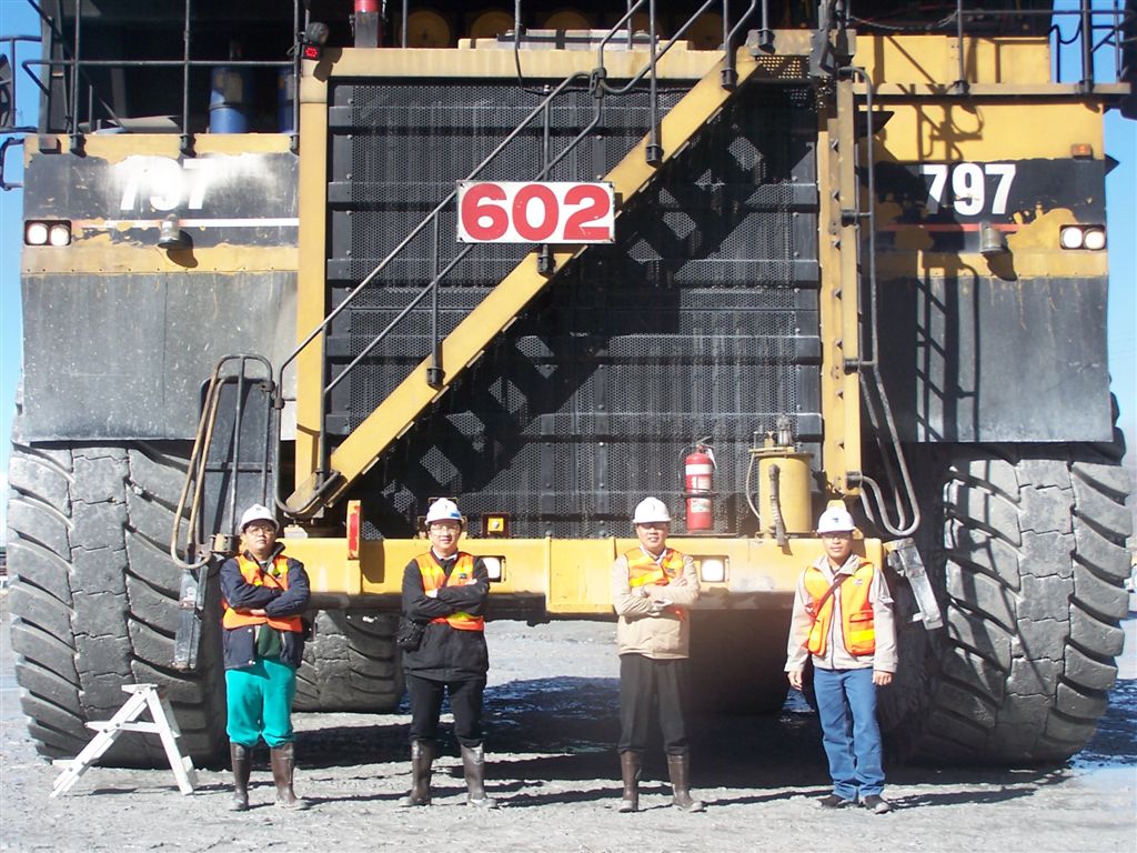 真實照片：一台Caterpillar 797超大型礦用卡車車尾，車身標示797字樣，四名戴安全帽與反光背心的礦場工作人員站在巨大輪胎前方，凸顯車輛尺寸